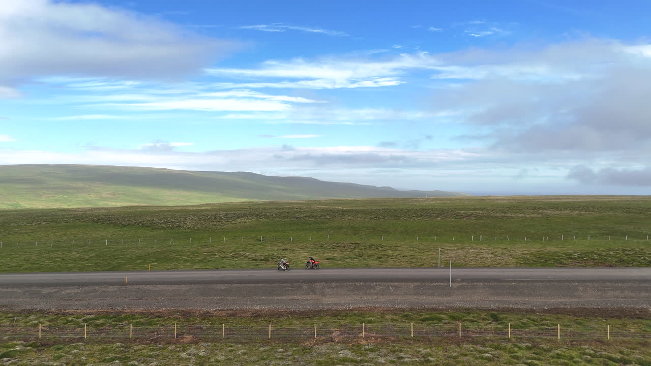 Luftfoto af to cyklister, der cykler på en afsidesliggende islandsk vej gennem åbne, grønne marker under en bred, blå himmel, der repræsenterer frihed, eventyrrejser og en bæredygtig udendørs livsstil.