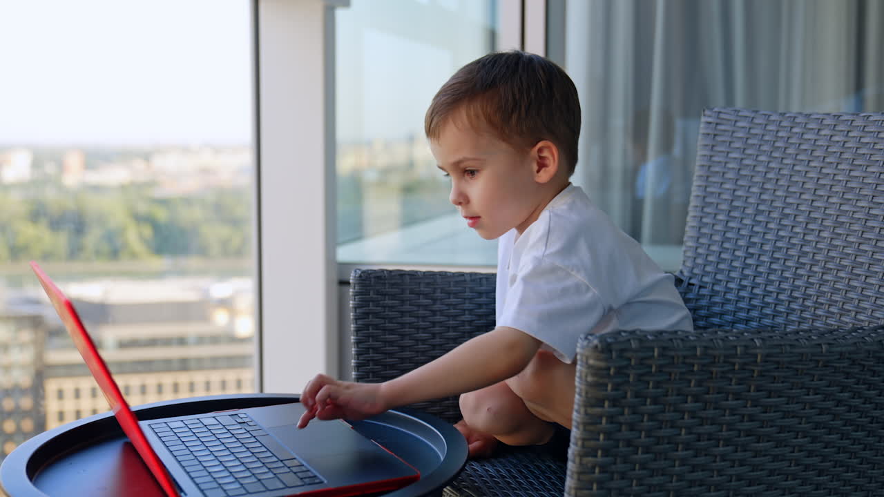 Boy watching laptop screen on sunny balcony. Child sitting cross-legged on modern balcony looking at laptop screen with sunlight and cityscape