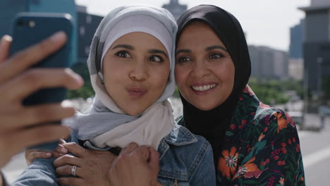 close up portrait of muslim mother and daughter smiling cheerful embrace posing taking selfie photo using smartphone in sunny urban city