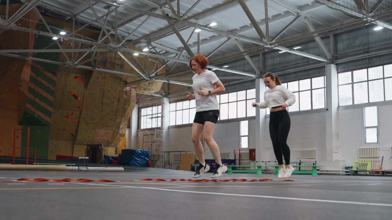 Exercícios de trabalho de pés de badminton em pavilhão desportivo coberto, treinador guia atleta através de padrões de passos em quadra marcada com teto alto e parede de escalada ao fundo, ênfase em passos laterais rápidos, equilíbrio