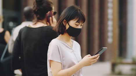 asian woman in mask standing on street and using smartphone