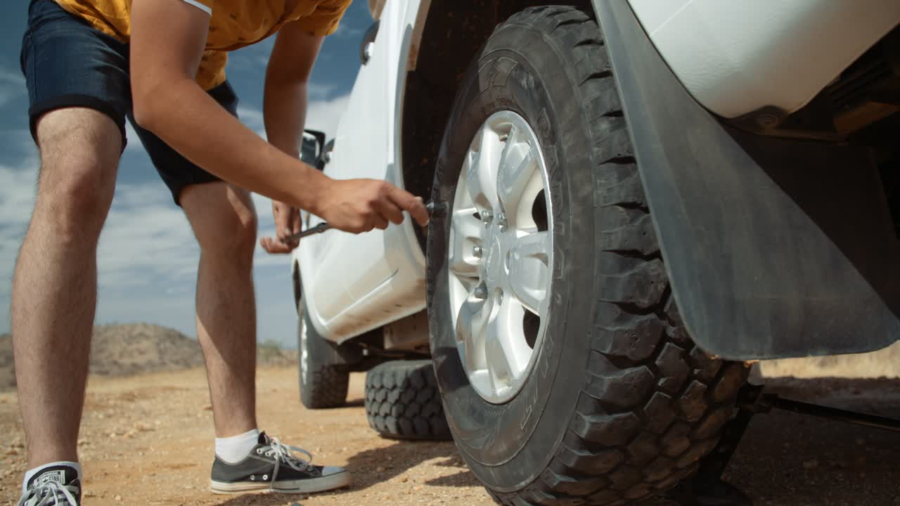 Static low wide hand held shot of a Caucasian male tourist in Africa as he tightens lug nuts with a lug wrench on a spare tire of a off-road vehicle after having a flat tire