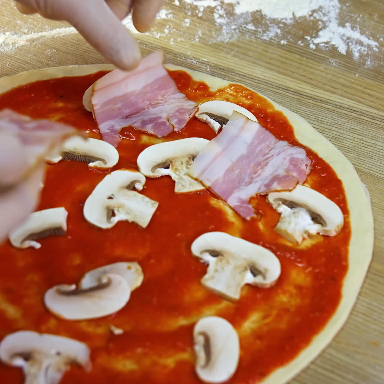 Putting mushrooms and bakon on pizza dough with tomato souce. Food video footage. Hands of a chef with ingredients for pizza