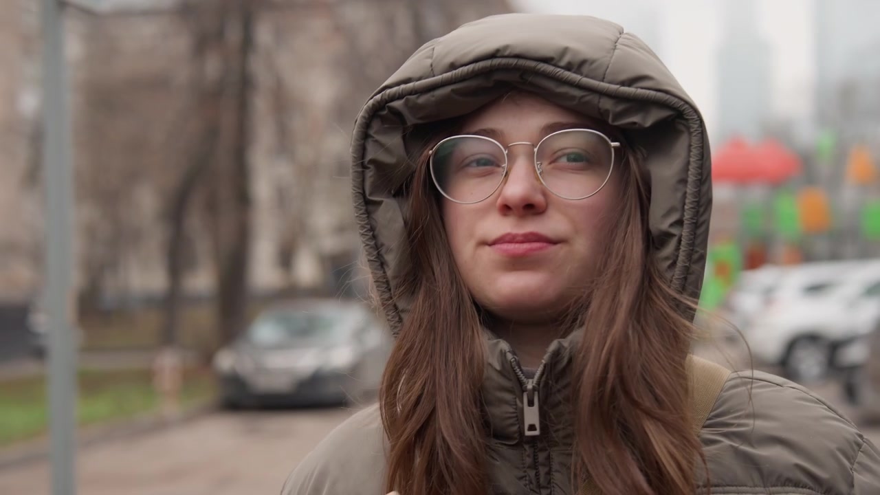 Giovane donna caucasica che si sistema il cappuccio all'aperto su un freddo marciapiede cittadino, che indossa occhiali e un piumino, leggere raffiche di neve le spolverano i capelli, le mani si muovono per proteggere il viso, le espressioni che cambiano da pensierose.