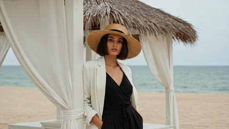 elegance on the shore: a stylish woman in a black dress and wide-brim hat poses at a beachside gazebo with a tranquil ocean backdrop