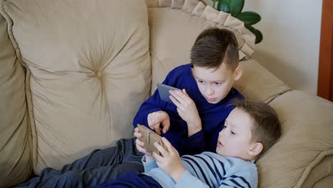 two cute boys relaxing on the couch and playing on his smaptphones at home. children spending time together holding phones in hands and playing competitive video games.