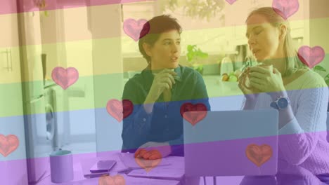 Animation-of-heart-emojis-and-rainbow-flag-over-caucasian-female-couple-using-laptop