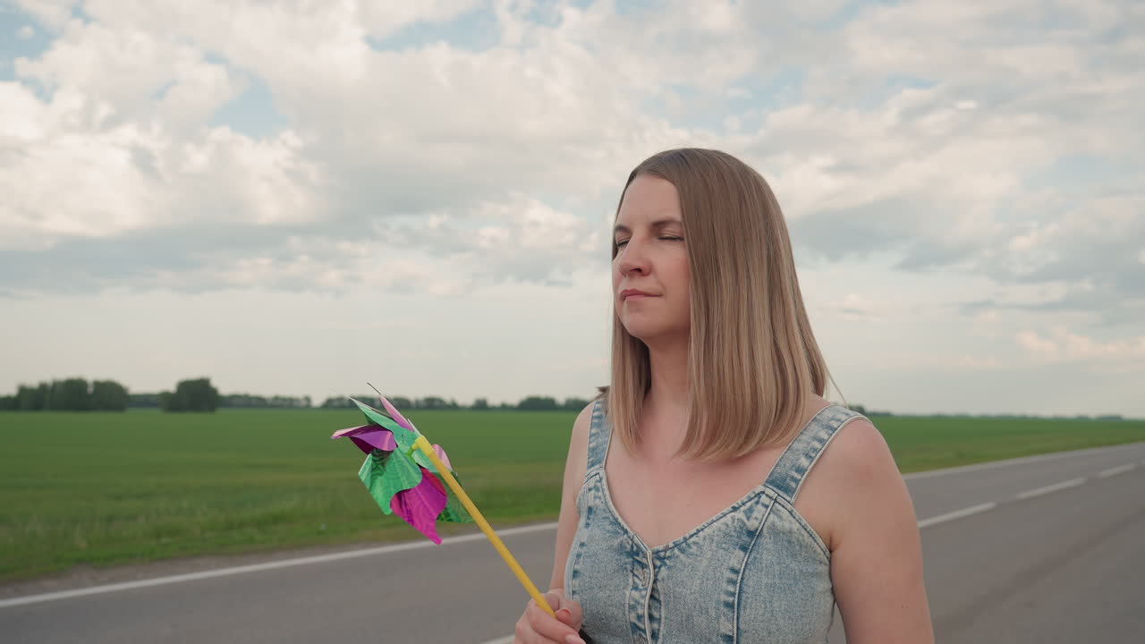Thoughtful young woman in denim dress walking along rural asphalt road holding vibrant pinwheel rotating gently in summer breeze beside endless green field under soft cloudy sky