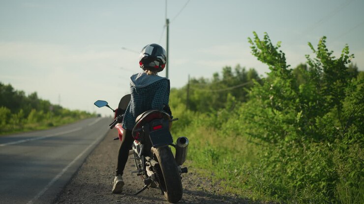 A back view of a female biker, relaxing on her bike, contemplatively looking to the left and right on a serene road with lush greenery and electric poles in the background