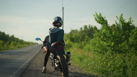 a back view of a female biker, relaxing on her bike, contemplatively looking to the left and right on a serene road with lush greenery and electric poles in the background