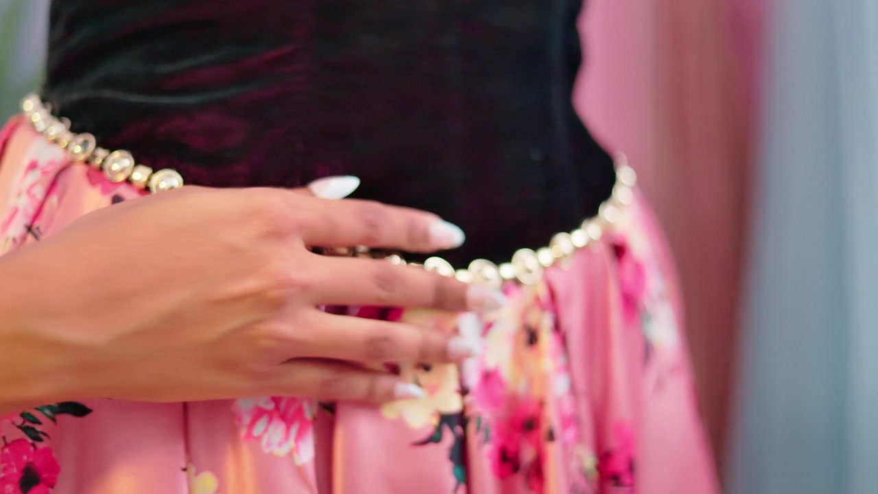 Bidikan jarak dekat tangan wanita yang menunjukkan detail ikat pinggang manik-manik hias pada pakaian bermotif bunga yang cerah di dalam studio pakaian