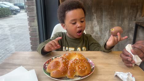 expressive and sweet 3-year-old black baby eating a chocolate egg and a cake in a cafeteria seated next to his mother