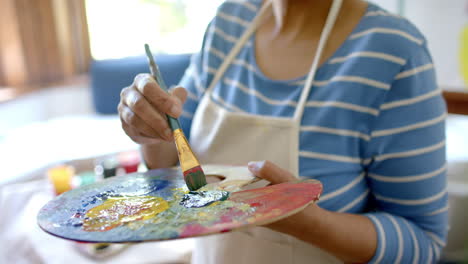 retrato de una feliz mujer biracial mezclando colores en la paleta en casa, en cámara lenta