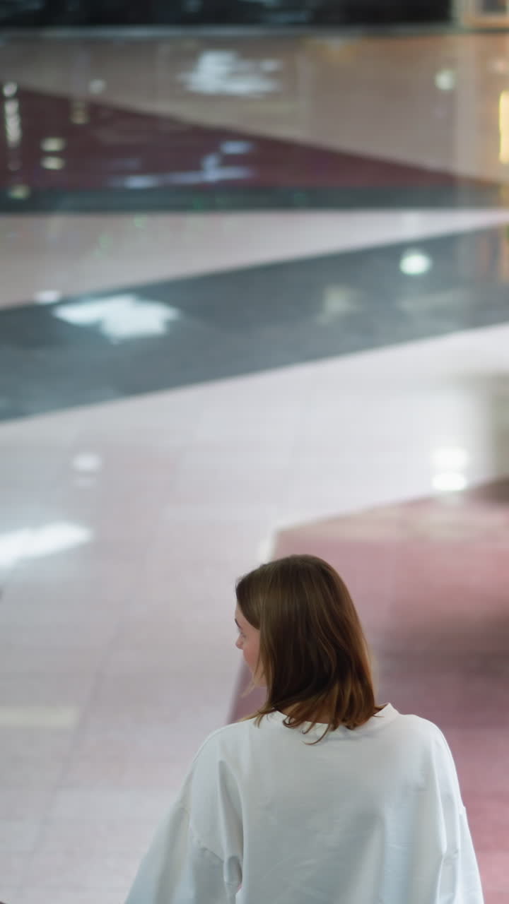 Back view of young woman on moving escalator in brightly lit indoor shopping mall, showcasing polished floor patterns, modern architectural details with partial view of people in the down floor