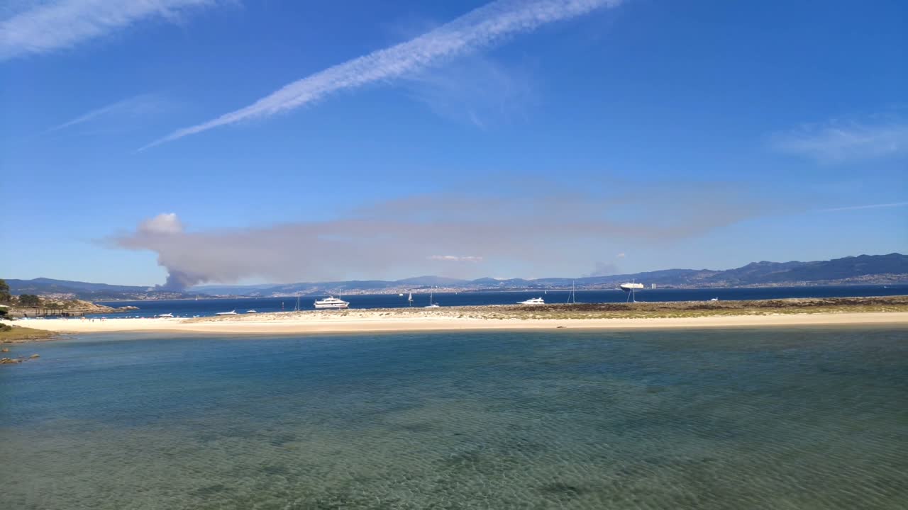 fuego en el horizonte detrás de la costa y la playa con yates de lujo y barcos anclados en un día soleado de verano, tiro panorámico girando a la derecha, islas cíes, pontevedra, galicia, españa