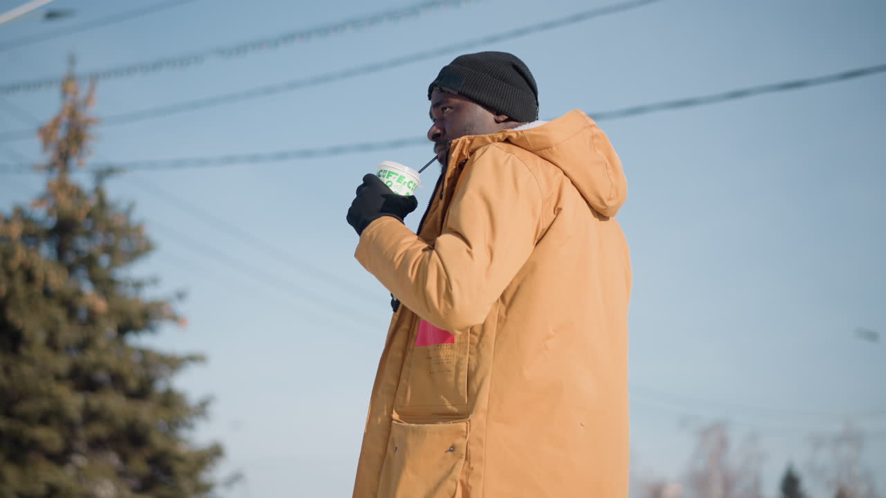 low side view of male artist holding drink cup in one hand holding camera in other while looking around busy snowy city under bright sun wearing gloves bag strap blur electricity wires