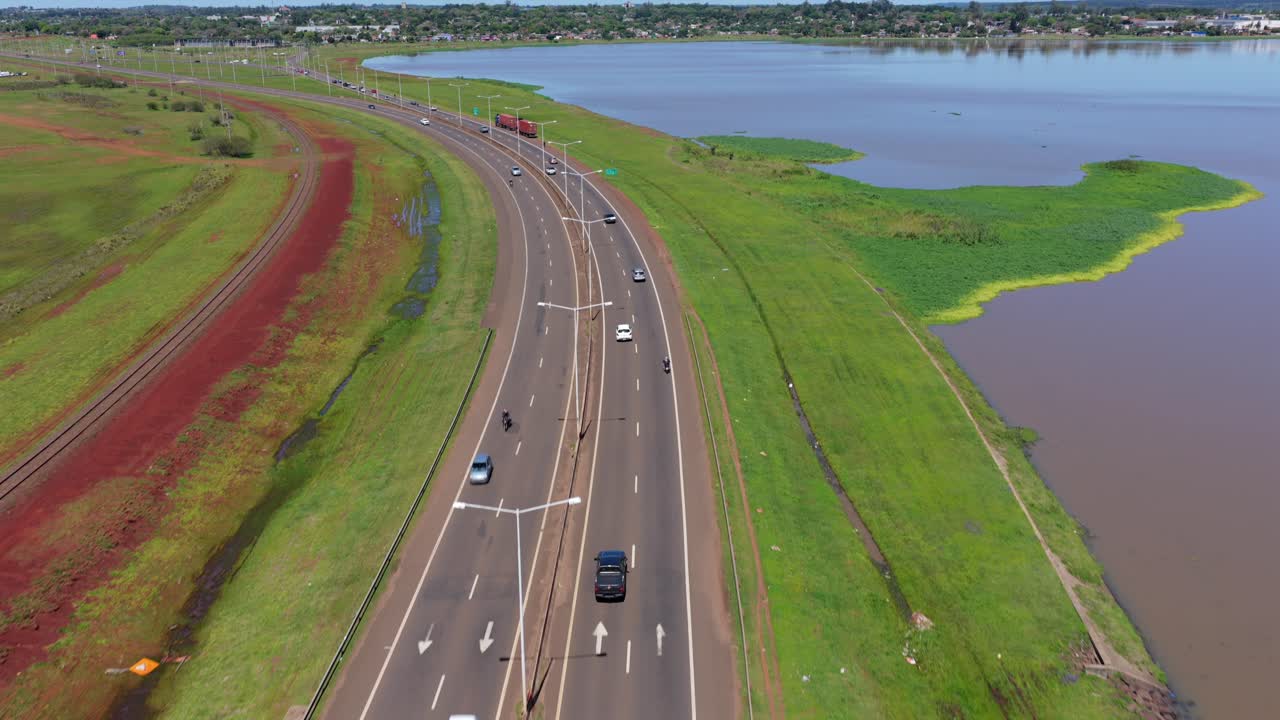 Drone tracks a car driving along Acceso Sur, Posadas, Misiones, Argentina, bordered by green landscape and the Paraná River on a clear day