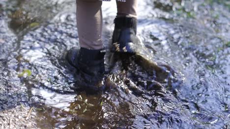 close up of toddler walking in a mud puddle with her black winter boots, splashing in the autumn forest