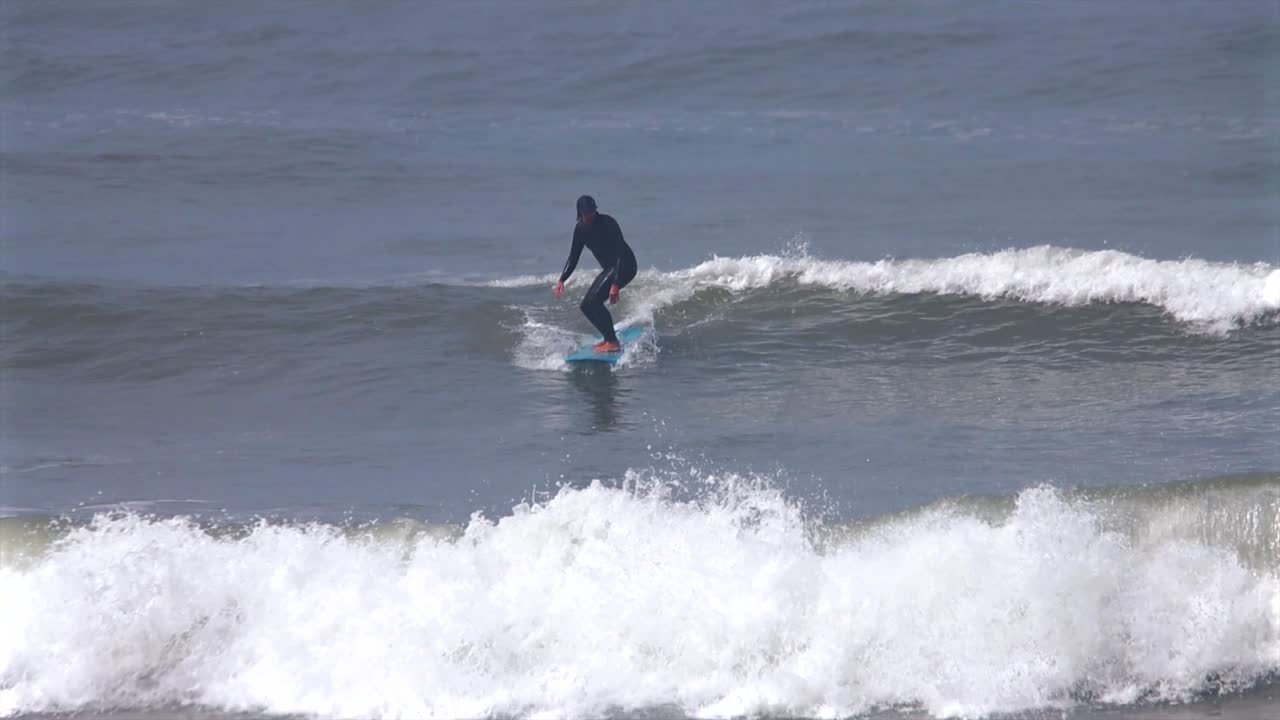 Surfista su longboard con muta che surfa una grande onda vetrosa a sinistra a Miraflores, Playa Makaha Beach, costa di Lima, Perù, Barranquito Barranca, giornata di sole, scuola di surf, teleobiettivo con zoom, panoramica a destra
