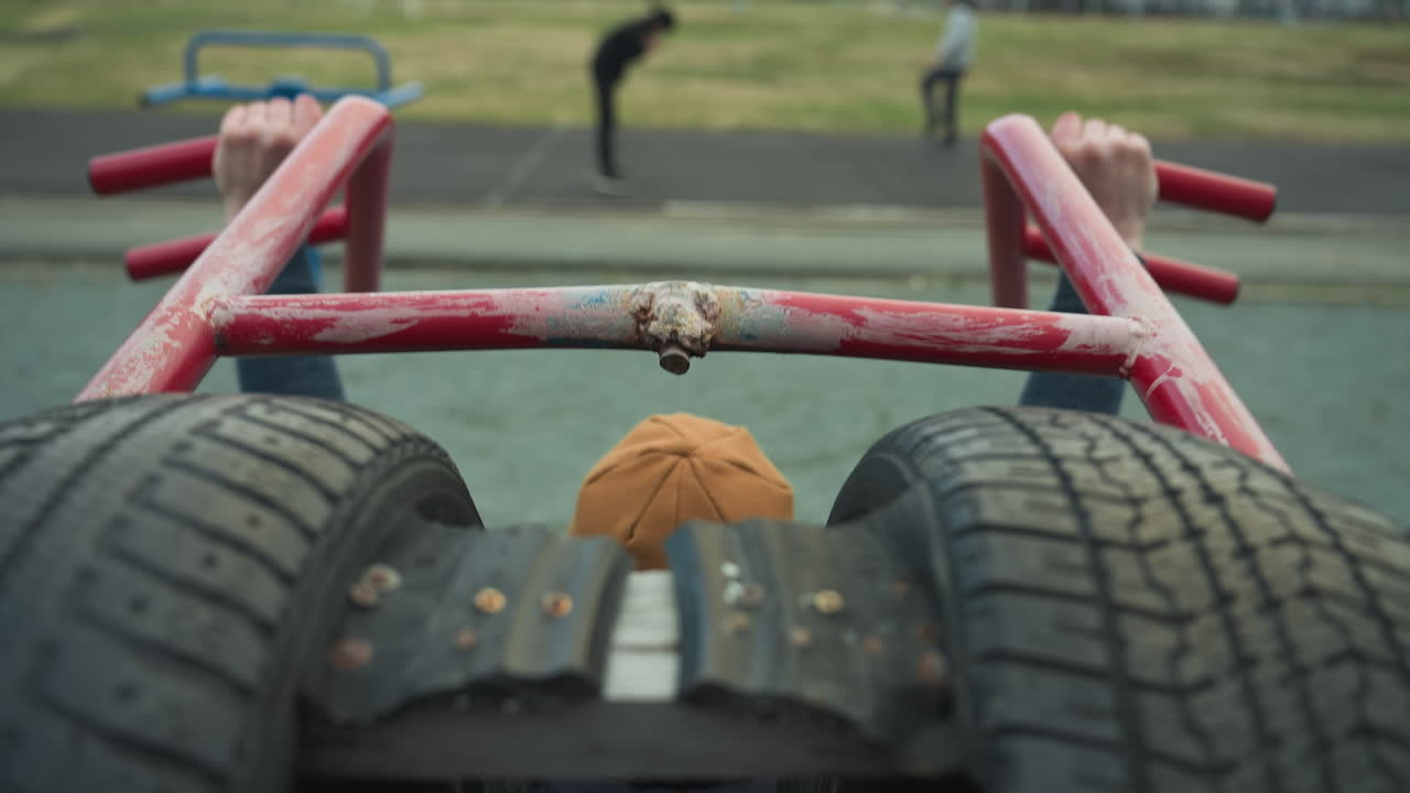 Back view of a coach doing pull-ups on workout equipment, pausing to rest, with a blurred view of two boys working out on a track with distant view of people on the field in the background
