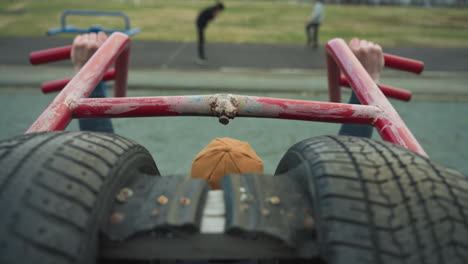 back view of a coach doing pull-ups on workout equipment, pausing to rest, with a blurred view of two boys working out on a track with distant view of people on the field in the background