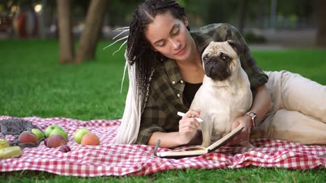 chica elegante acostada en cuadros en el césped en un parque y haciendo notas mientras un pequeño pug sentado a su lado