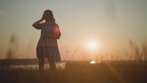 woman in a blue gown, using her smartphone and headset, enjoys a radiant sunset in a lush field, gently swaying to the music