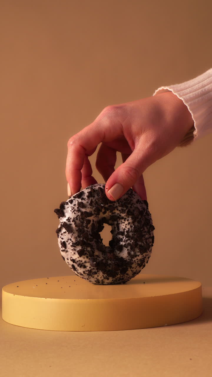 Hand spins chocolate donut with white icing and dark cookie crumbs in place on round pedestal. Warm beige studio background with clean minimal composition
