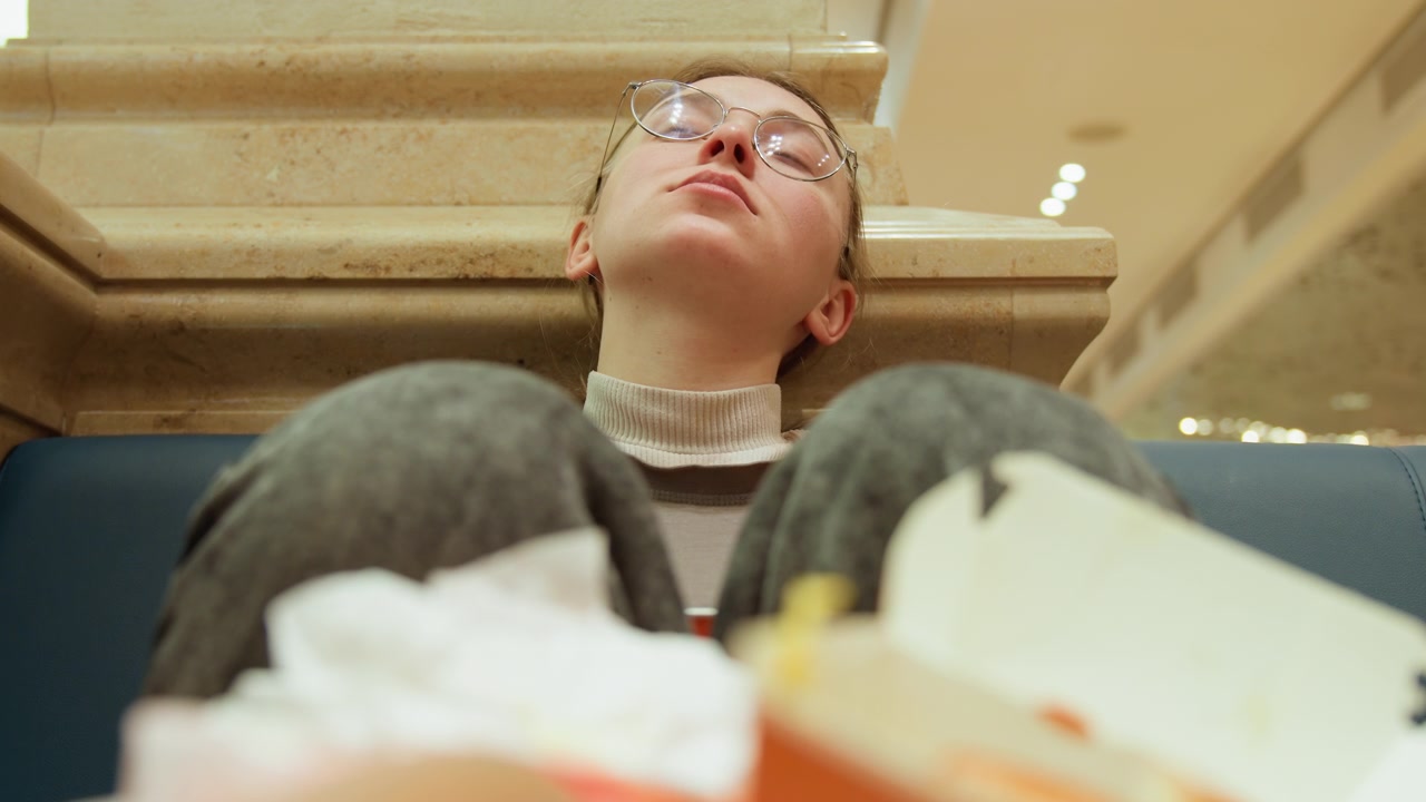 Studente esausto accasciato su una panchina nell'area ristorazione di un centro commerciale, occhiali che riflettono le luci dall'alto, contenitori da asporto e tovaglioli accartocciati in primo piano, illuminazione ambientale calda, ritratto spontaneo.