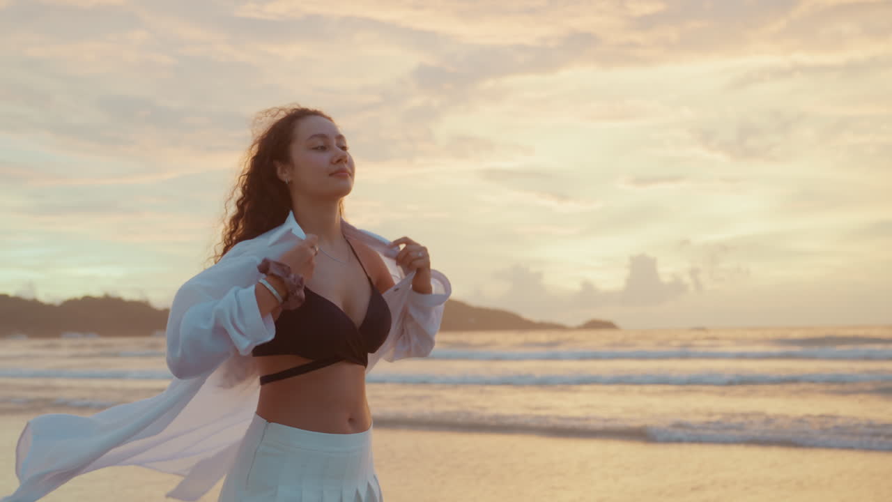 Donna sulla passeggiata lungomare all'ora d'oro. Una donna con i capelli ricci osserva l'orizzonte al tramonto sulla costa. Una figura femminile in abito bianco guarda in lontananza lungo la tranquilla riva al crepuscolo.