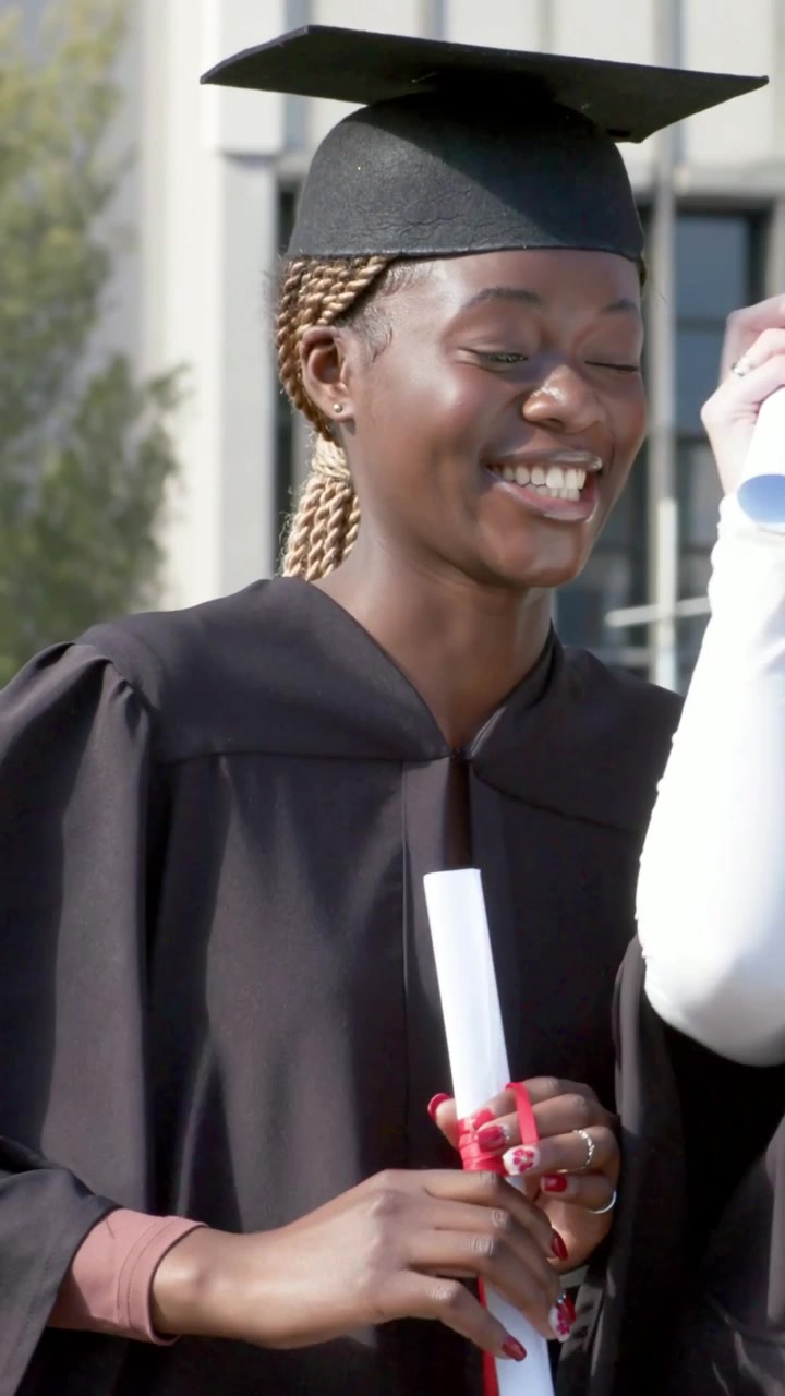 Vertical video: Laughing grad taking diploma from peer on campus in cap and black gown, celebrating