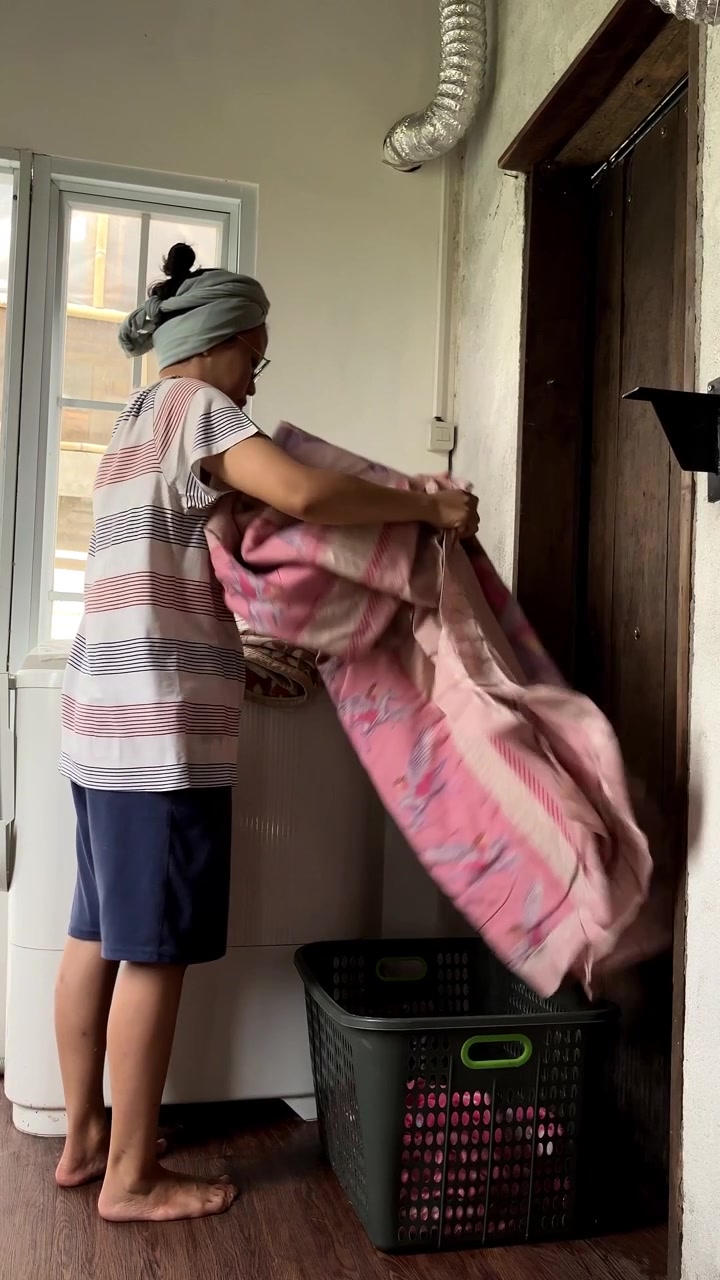 Slow motion vertical video of an Asian woman lifting a large heavy blanket toa plastic basket. She is doing her daily household laundry chores in a rustic indoor setting.