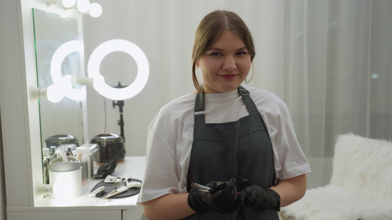 Beautician in apron and gloves looks at camera while holding makeup brushes beside table filled with professional cosmetic tools. Vanity mirror and ring light enhance modern salon atmosphere