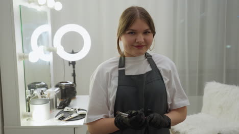 beautician in apron and gloves looks at camera while holding makeup brushes beside table filled with professional cosmetic tools. vanity mirror and ring light enhance modern salon atmosphere