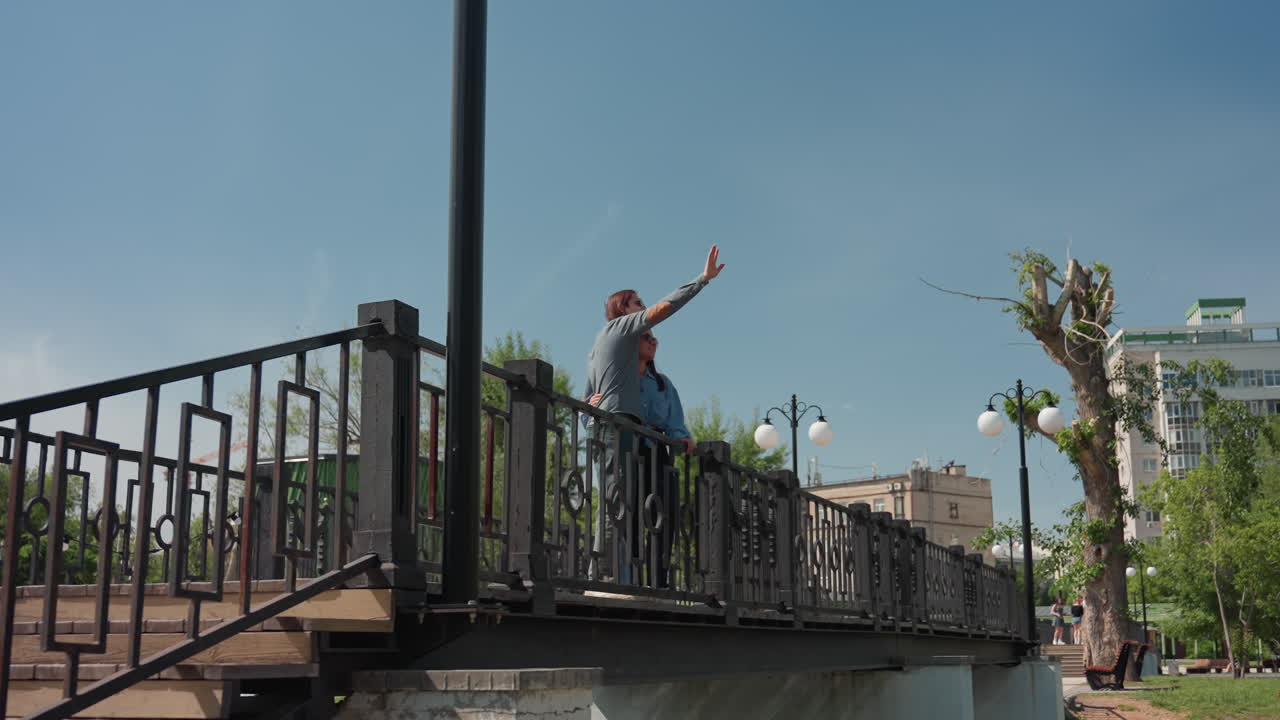 woman indicates skyline confidently upward, female figure signals horizon with assured gesture, an elegant woman points toward distant city skyline from ornate bridge structure with assuredness