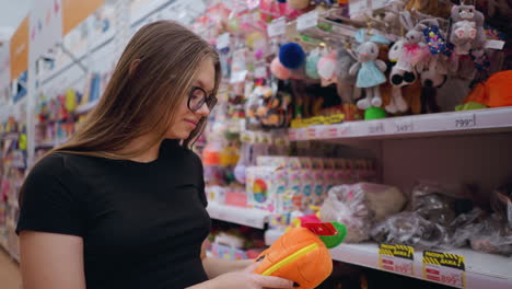 young woman in black t-shirt and jeans browsing toy store shelves, reaching for soft plush toy, vibrant stuffed animals and keychains hang above as she observes item