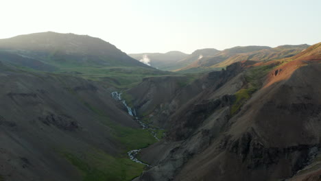 Birds-eye-flying-towards-amazing-iceland-panorama-with-river-flowing,-waterfall-and-moss-covering-mountains.-Aerial-drone-view-of-breathtaking-scandinavian-landscape-at-sunset.-Beauty-in-nature