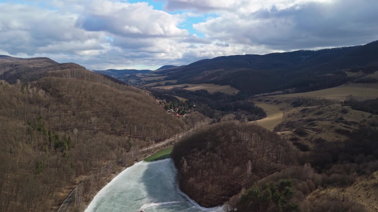 In Slovakia's Košice Region, the frozen Ružín reservoir lies among Carpathian hills and forested valleys under an overcast winter sky in an aerial view.