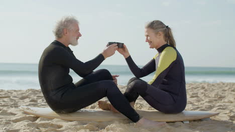 senior couple in wetsuit drinking tea and talking together while sitting on a surfboard at the beach