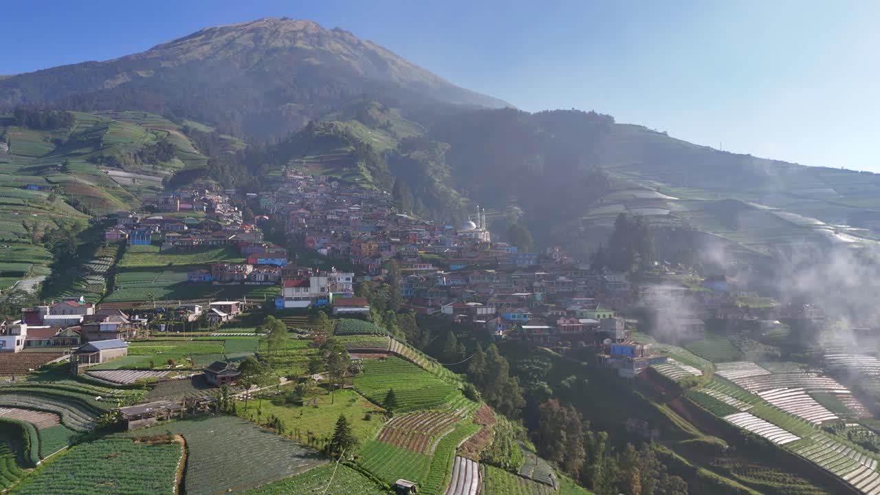 Aerial view of scenic mountain village surrounded by lush green terraced farms. Morning mist and clear blue skies create a peaceful rural atmosphere. Nepal van Java, Indonesia