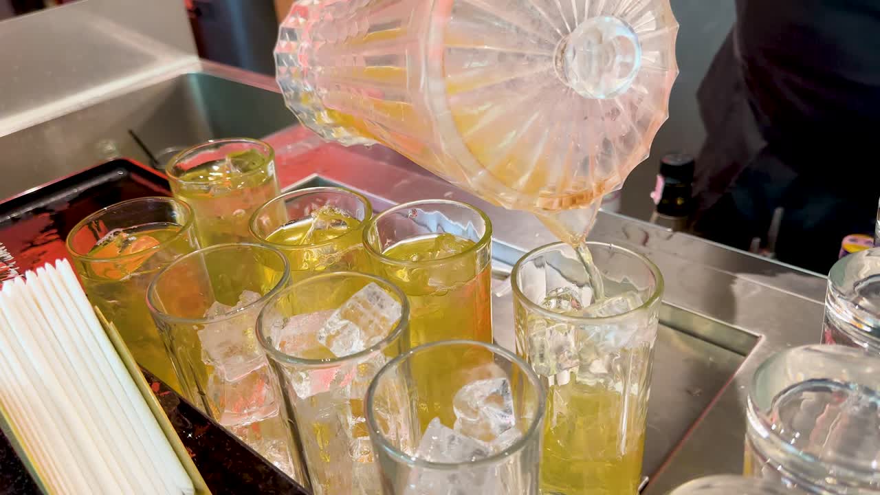 Bartender pours cold iced tea from glass pitcher into ice-filled glasses on counter
