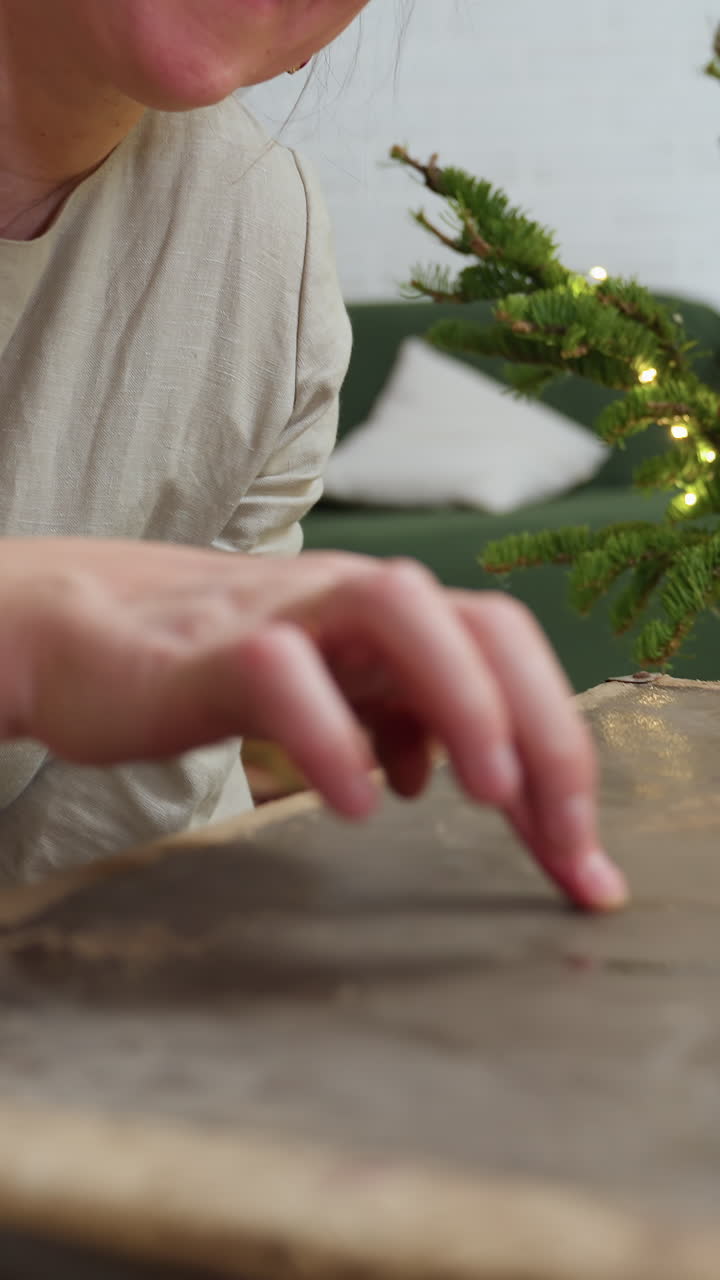Mujer caucásica envolviendo regalos en el interior, vestido de lino, primer plano de manos alisando papel de manualidades, pequeño árbol de Navidad iluminado al fondo, anfitriona sonriente organizando una decoración festiva, ambiente acogedor.