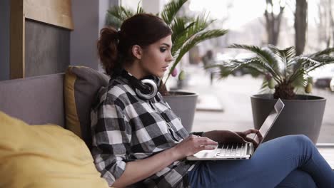 mujer joven concentrada, sentada en la habitación o espacio de trabajo con ventanas. trabajando en una computadora portátil en sus piernas y auriculares alrededor del cuello. espacio moderno y ligero. vista lateral