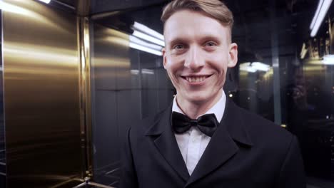 young man in a suit with bow tie rides in the elevator looking in the camera and smiling. evening event.