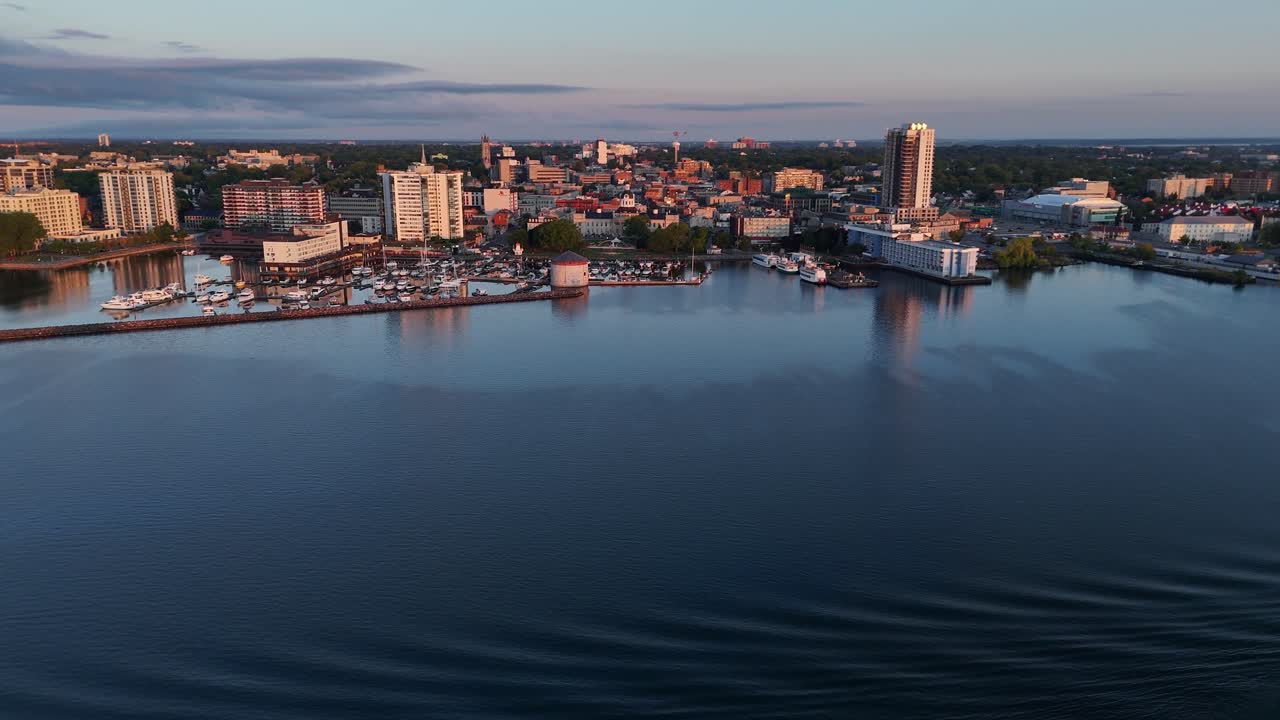 Cinematic 4K drone footage of Kingston, Ontario, Canada at golden hour. The aerial view captures the Wolfe Island Ferry crossing the waterfront with the city’s historic City Hall and downtown skyline