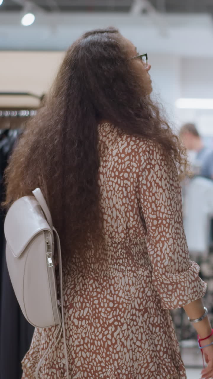 mujer con cabello largo y rizado y vestido elegante navegando por los estantes de ropa en la tienda, llevando una mochila blanca, otros compradores en el fondo, creando una animada atmósfera comercial con iluminación suave