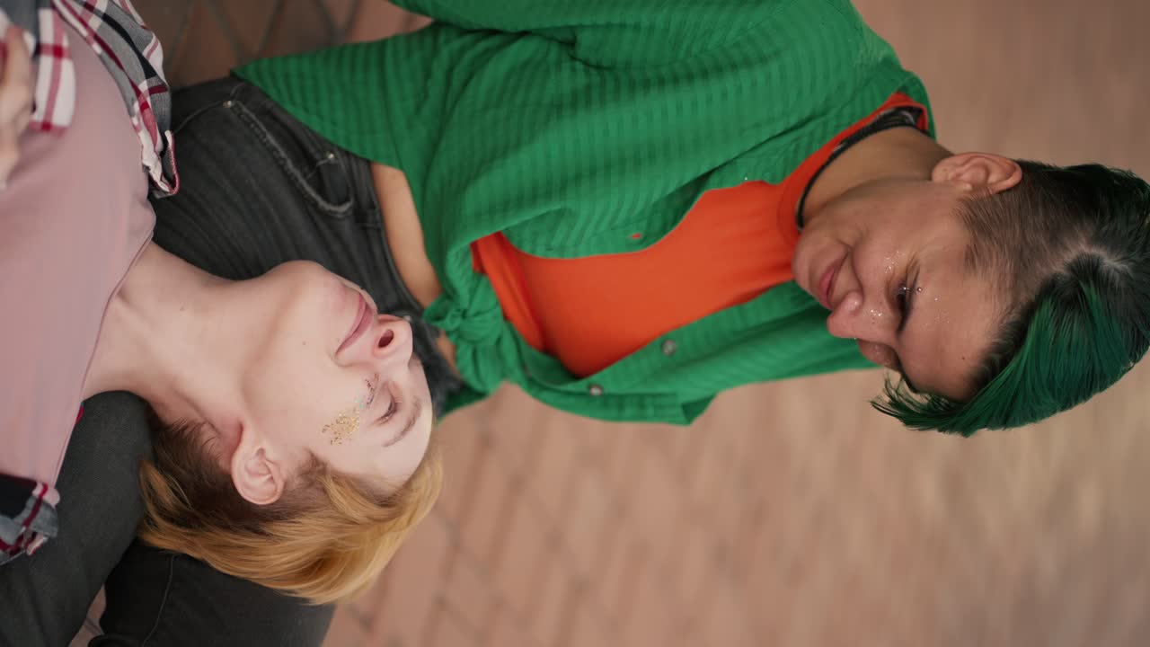 вертикальное видео: блондинка с короткой стрижкой в розовой клетчатой рубашке лежит на коленях своей девушки в зеленой рубашке с короткой зеленой стрижкой во время ее свидания в городе