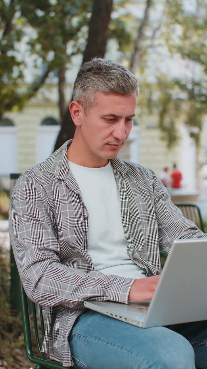 Happy greyhaired man opens laptop and working on project sitting on chair in city street outdoors