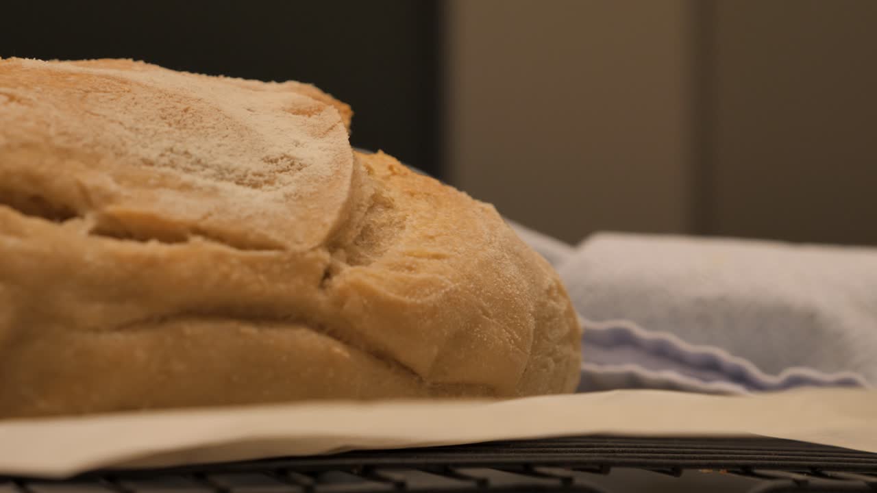 Slow motion pan through to freshly baked loaf of sour dough bread topped with flour sitting on kitchen bench with tea towel and tray, low depth of field