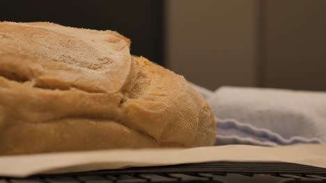Slow-motion-pan-through-to-freshly-baked-loaf-of-sour-dough-bread-topped-with-flour-sitting-on-kitchen-bench-with-tea-towel-and-tray,-low-depth-of-field